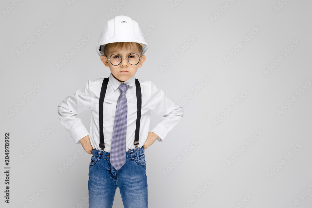 A charming boy in a white shirt, suspenders, a tie and light jeans stands on a gray background. Boy with a white helmet A charming boy in a white shirt, suspenders, a tie and light jeans stands on a gray background. Boy with a white helmet