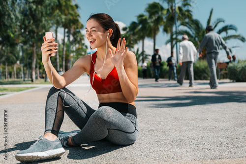 Hi. Portrait of joyful girl is having online communication. She is waving hand to cellphone and smiling. Woman is sitting on alley outdoor and relaxing after workout