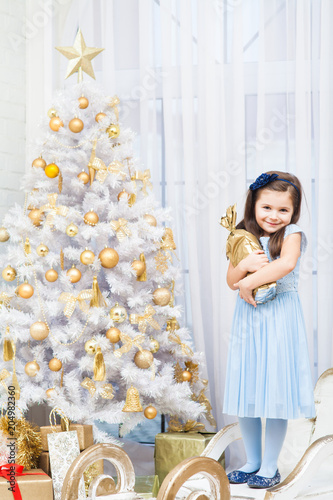 smiling girl in a blue dress and blue shoes stands near a white Christmas tree