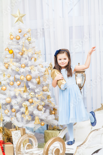 smiling girl in a blue dress and blue shoes stands on one leg near a white Christmas tree