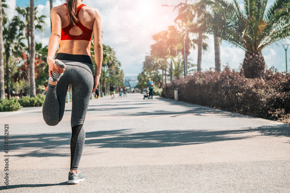 Sport is my life. Focus on woman back warming up her leg before running. She is standing on alley near tropic palms. Copy space