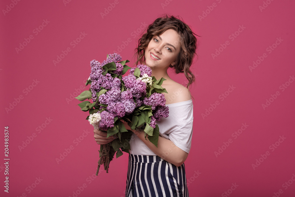 A young slender girl in stylish clothes is holding a bouquet of purple flowers. She is surprised at the gift she was given on a date. On a pink background with space for text A young slender girl in stylish clothes is holding a bouquet of purple flowers. She is surprised at the gift she was given on a date. On a pink background with space for text