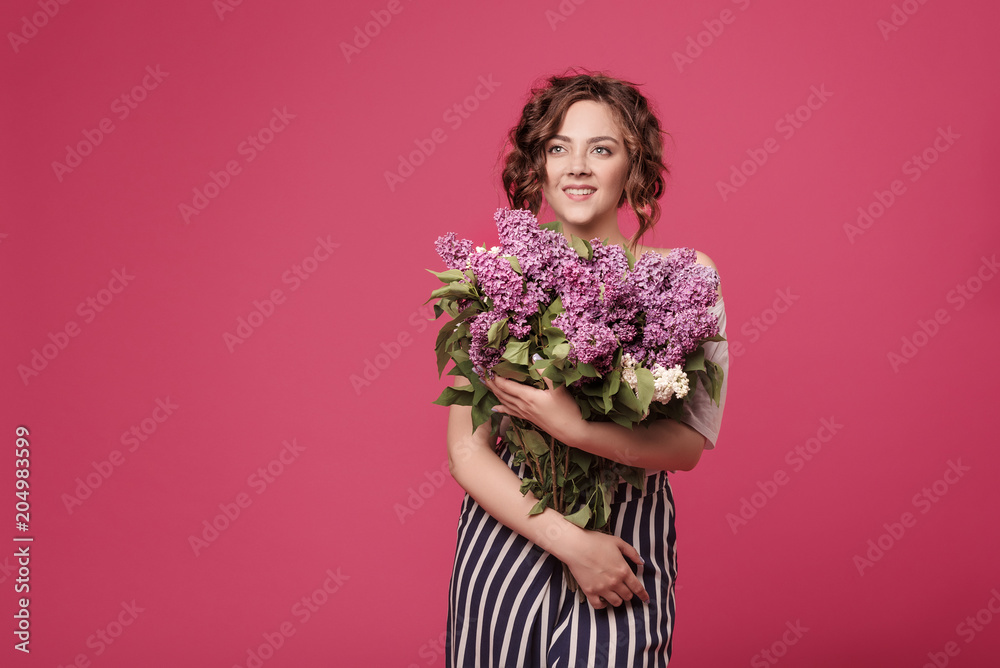 A young slender girl in stylish clothes is holding a bouquet of purple flowers. She is surprised at the gift she was given on a date. On a pink background with space for text A young slender girl in stylish clothes is holding a bouquet of purple flowers. She is surprised at the gift she was given on a date. On a pink background with space for text