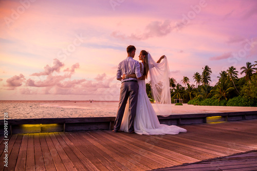 Beautiful married couple standing on a jetty on Maldives watching amazing purple blue sunset. Tropical wedding. photo