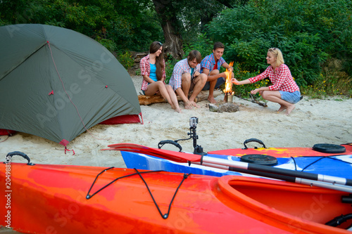 Young Happy Travelers Resting at Evening with Fire on the Sand Beach near Kayaks and Tent.