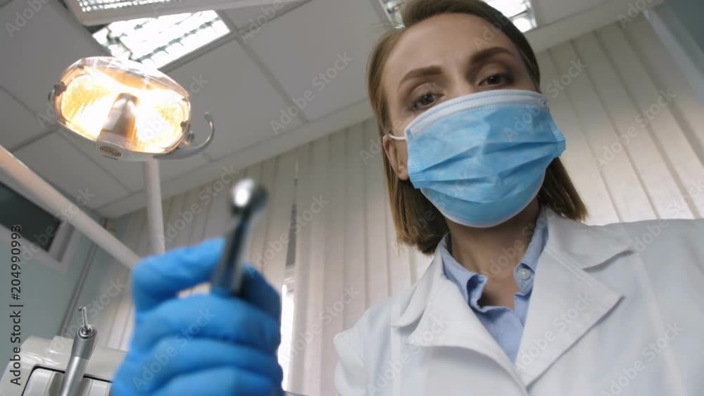 Beautiful young female dental doctor putting on face mask, taking dental drill machine and mirror and starts treating patient's tooth decay. Low angle shot, patient point of view