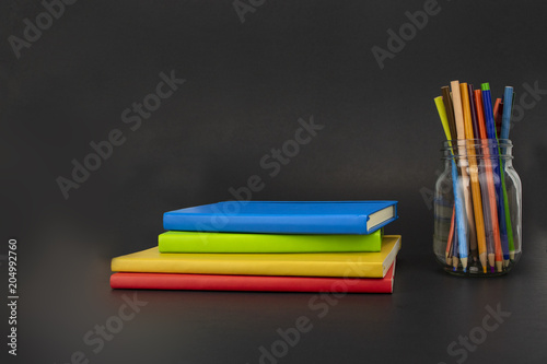 A set of colorful notebooks sits next to a set of bright colored pencils in a jar. Room for text! Beautiful black background! 