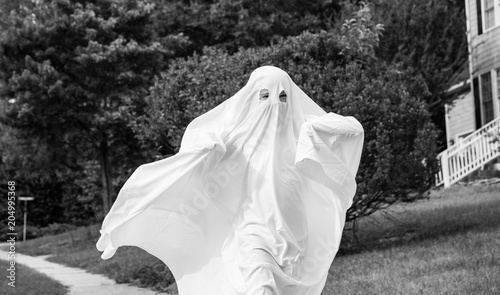 Fotografie A black and white photo of a child in a ghost costume made out of a bedsheet running down the sidewalk