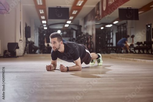 one young man, looking away, plank exercise, gym floor, unrecognizable people behind (out of focus).