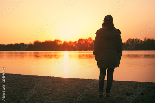 Fototapeta Naklejka Na Ścianę i Meble -  a young caucasian woman standing near the shoreline of a lake watching the sunset by herself thinking and contemplating