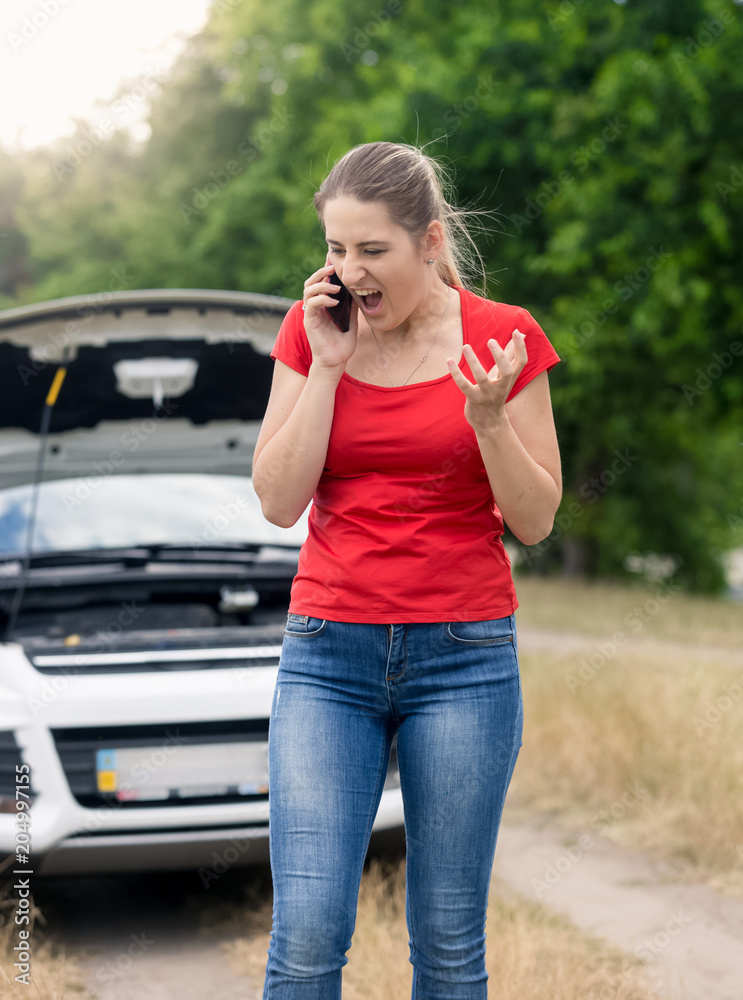 Angry young woman standing at broken car in field and shouting in mobile phone Angry young woman standing at broken car in field and shouting in mobile phone