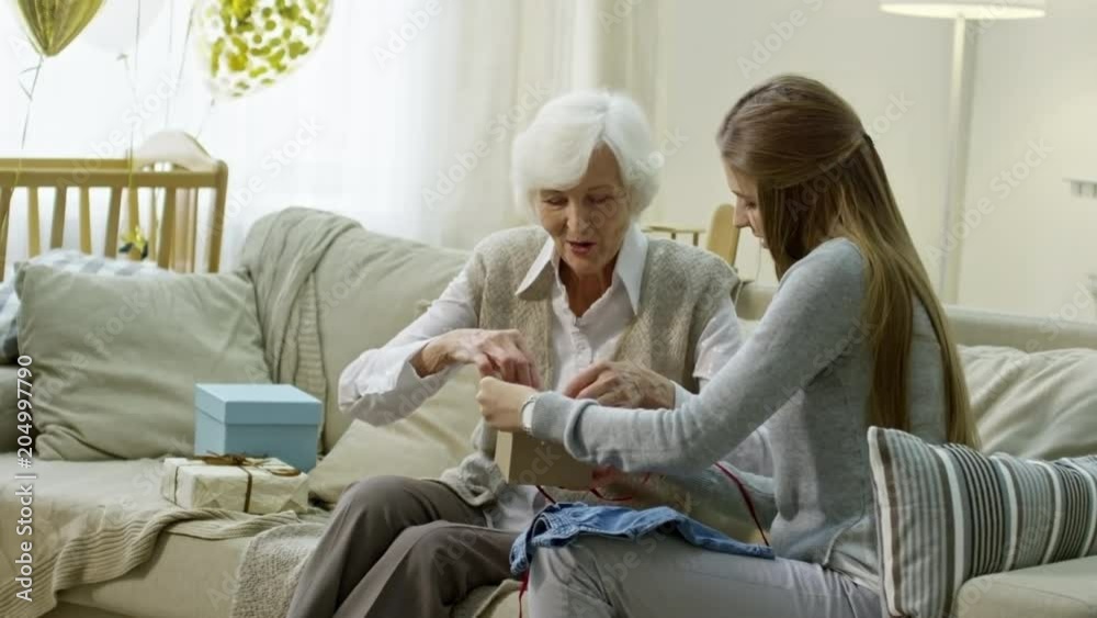 Medium shot of happy grandmother and young mother sitting on sofa and opening presents with clothes for baby Medium shot of happy grandmother and young mother sitting on sofa and opening presents with clothes for baby