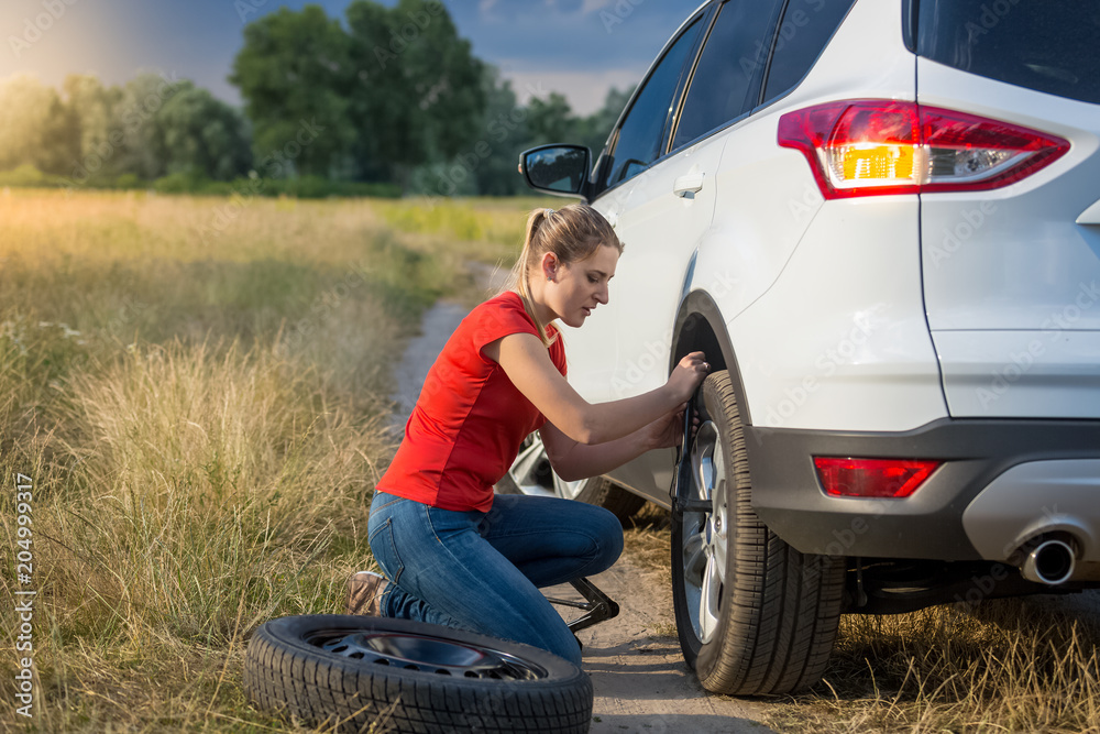 Upset young woman unscrewing wheel nuts to change flat tyre on deserted countryside road Upset young woman unscrewing wheel nuts to change flat tyre on deserted countryside road