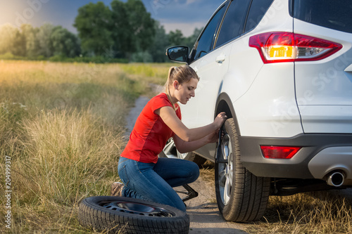 Upset young woman unscrewing wheel nuts to change flat tyre on deserted countryside road