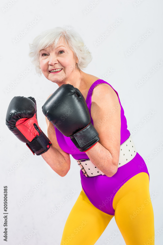 portrait of beautiful sporty senior lady in boxing gloves smiling at camera isolated on grey portrait of beautiful sporty senior lady in boxing gloves smiling at camera isolated on grey