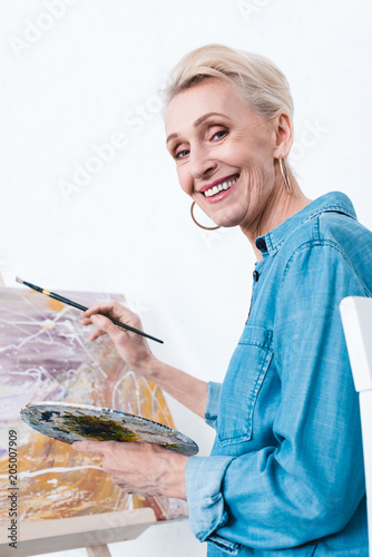 senior female artist with palette and brush painting in workshop