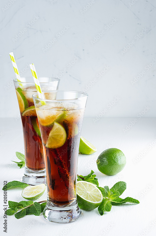 Refreshing alcohol beverage cuba libre with cola, ice, mint, lime, straw in two wet long glasses in modern white kitchen interior, copy space. Refreshing alcohol beverage cuba libre with cola, ice, mint, lime, straw in two wet long glasses in modern white kitchen interior, copy space.