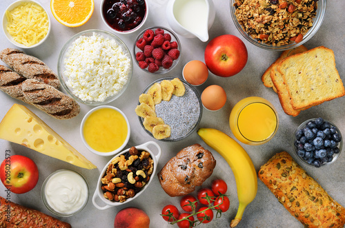 Healthy breakfast ingredients, food frame. Granola, egg, nuts, fruits, berries, toast, milk, yogurt, orange juice, cheese, banana, apple on gray background, top view, copy space.