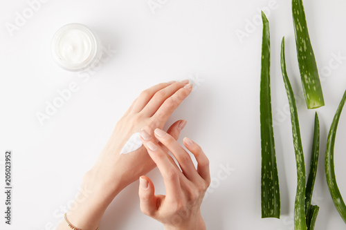 cropped image of woman applying cream on hands, aloe vera leaves and cream in container on white surface
