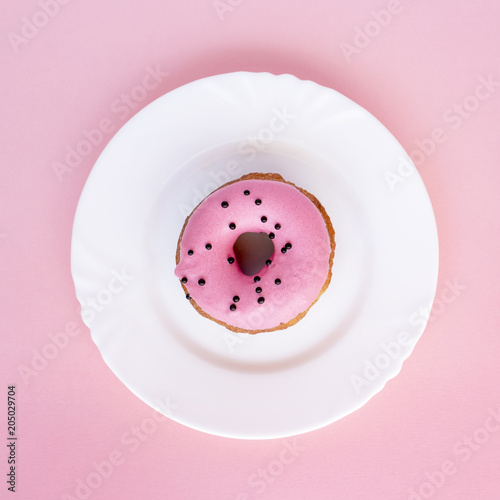 Donut covered with pink frosting and sprinkled with black roundels on white plate on pink background