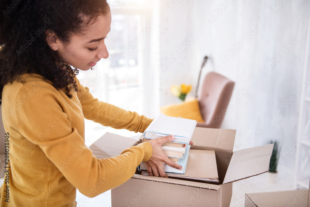 Necessary books. Charming curly-haired girl fitting a pile of books into the box while preparing to move out of an old flat Necessary books. Charming curly-haired girl fitting a pile of books into the box while preparing to move out of an old flat