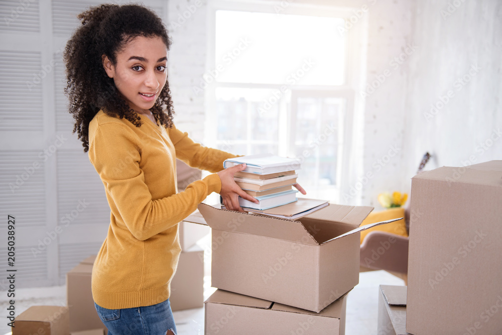 New start. Pleasant young girl taking a pile of books out of the box and posing for the camera while moving in to a new apartment