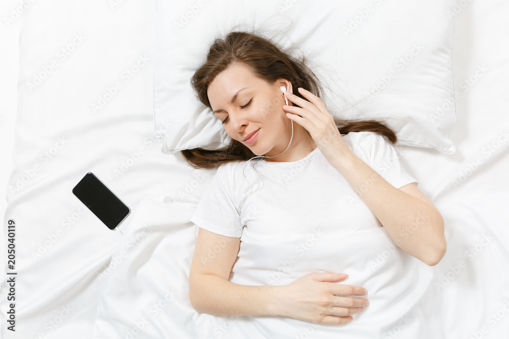 Top view of happy young woman lying in bed with white sheet, pillow, blanket, listen music from earphones in mobile phone. Calm beautiful female spending time in room. Rest, relax, good mood concept. Top view of happy young woman lying in bed with white sheet, pillow, blanket, listen music from earphones in mobile phone. Calm beautiful female spending time in room. Rest, relax, good mood concept.