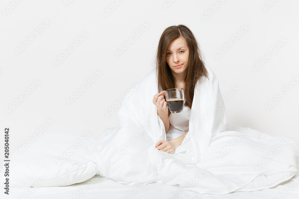 Young tired brunette woman sitting in bed with cup of coffee, white sheet, pillow, wrapping in blanket on white background. Female spending time in room. Rest, relax, good mood concept. Copy space.
