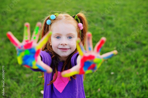 cheerful little girl with red hair shows her hands dirty with multicolored paints and smiles