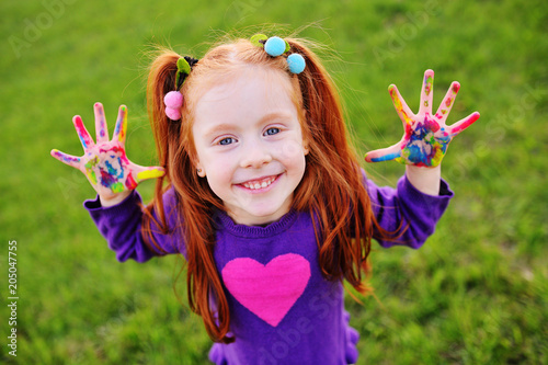 cheerful little girl with red hair shows her hands dirty with multicolored paints and smiles