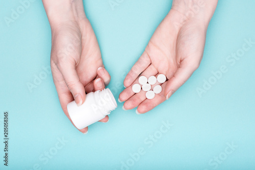 Close up of female hands holding medication bottle and white pills over pastel blue background. Patient taking medication.