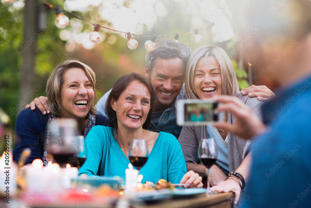 Obraz premium Some friends in their forties gathered around a table in the garden for a good time. A man takes a picture of a group of friends