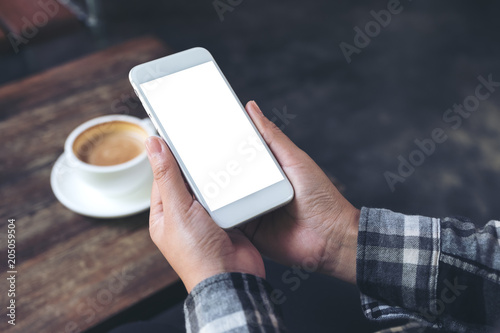 Mockup image of a woman's hand holding white mobile phone with blank desktop screen with coffee cup on wooden table in cafe
