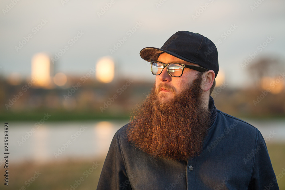Bearded man in real life. Portrait of young guy with large beard in glasses and a black cap at sunset in front of a river in summer. Bearded man in real life. Portrait of young guy with large beard in glasses and a black cap at sunset in front of a river in summer.