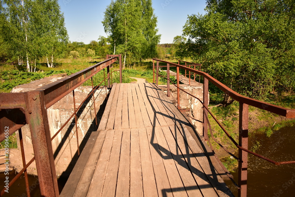 landscape wooden bridge over the river, rusty metal railings, background green trees, blue sky