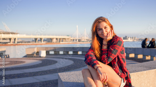 Beautiful young redheaded woman sitting on waterfront, enjoying nature and the good weather. The girl in red plaid shirt