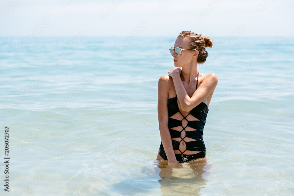 Pensive pretty girl in sunglasses and black swimsuit posing near the sea, looking away. Pensive pretty girl in sunglasses and black swimsuit posing near the sea, looking away.