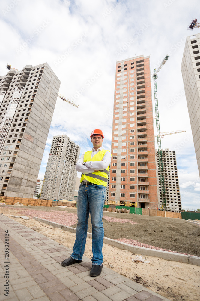 Confident male acrhitect in safety vest and red hardhat standing on building site
