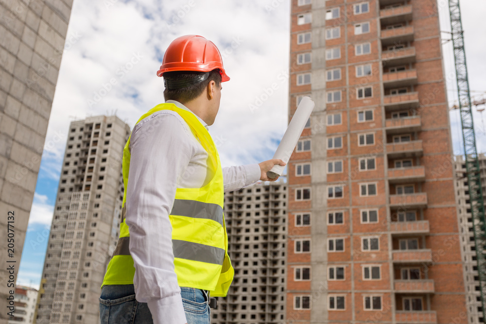 Male construction worker in hardhat holding blueprints and pointing at building under construction Male construction worker in hardhat holding blueprints and pointing at building under construction