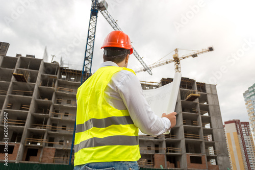 Rear view image of male architect in hardhat and vest looking at working building cranes