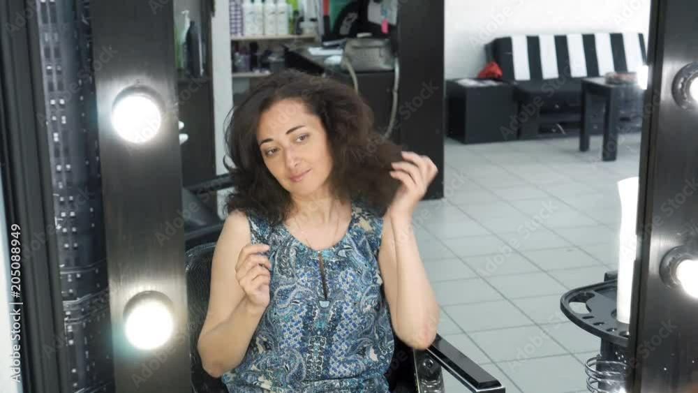 A woman with a beautiful hairstyle admires herself, sitting in front of a mirror in a hairdresser. Straightens her hair and looks in the mirror. The girl is preparing for an important event A woman with a beautiful hairstyle admires herself, sitting in front of a mirror in a hairdresser. Straightens her hair and looks in the mirror. The girl is preparing for an important event
