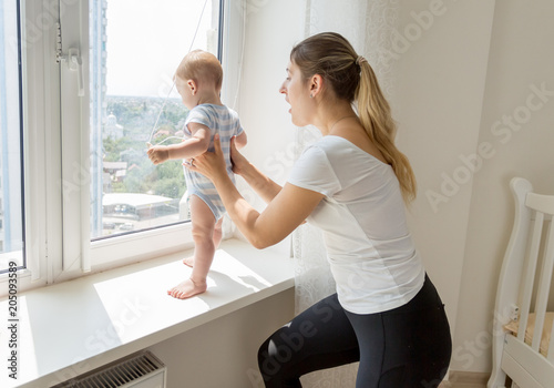 Young mother catching and holding her baby son standing on sindowsil and looking out of the window