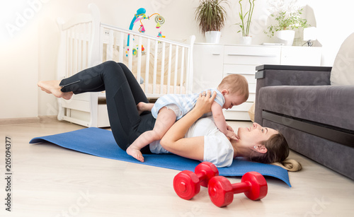 Cute baby boy sitting on young mother exercising on fitness mat at home