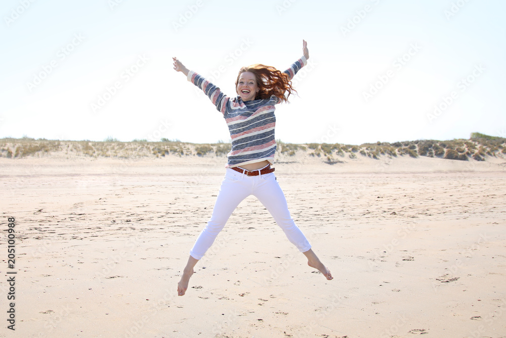 Obraz premium Hübsche rothaarige Frau an einem Strand springt vor Freude mit ausgebreiteten Armen in die Luft
