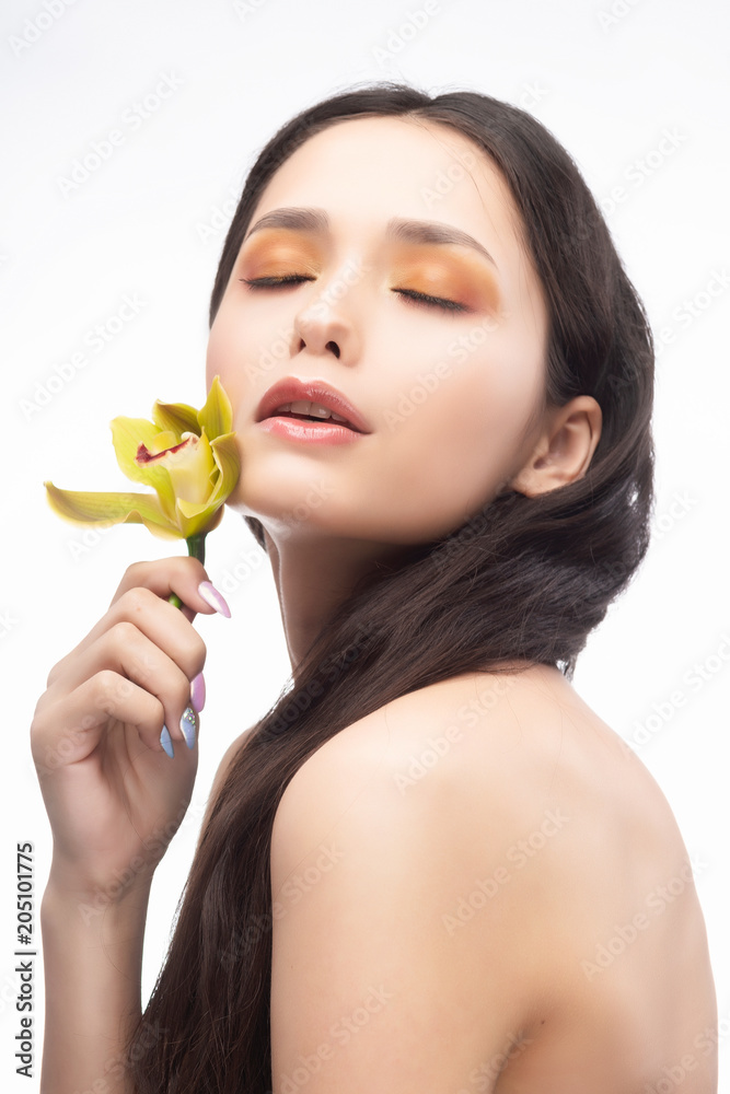 A beautiful brunette nude shoulders girl sensually holds a flower of an orchid in her hands. Nude make-up. Healthy smooth skin