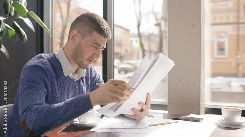 Young male employee is in working process with contract, sitting at table in cafe, businessman is holding, reading documents on background window, papers and telephone is on desk. Business person