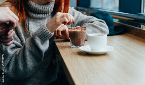 a girl in a cafe stirs, eating a hot chocolate spoon. the guy opens the sugar for coffee, tea in a white mug that is on the table.