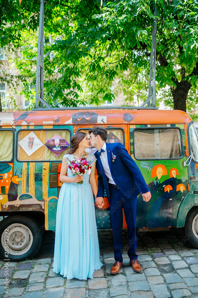 Funny newlyweds stay on background old painty bus. Gorgeous bride in blue wedding dress with bouquet hugging with handsome groom in blue stylish suit, newlywed couple portrait outdoors.