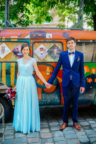 Funny newlyweds stay on background old painty bus. Gorgeous bride in blue wedding dress with bouquet hugging with handsome groom in blue stylish suit, newlywed couple portrait outdoors.