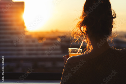 Silhouette of girl with a glass of juice at sunset. View on the town from above